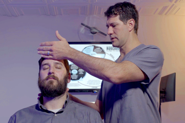 chiropractor hypnotherapist Dr. Theo Kousouli stands to the side of a male patient with a beard who is seated and performs a cervical spine palpation exam of the patient's neck. A television is behind them both displaying the Kousouli Method used by the doctor for working with his patients.